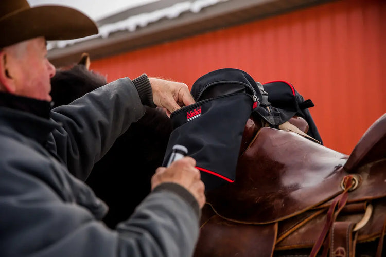 Man putting a beverage inside horn saddlebag the riding saddle on a horse in front of a red barn