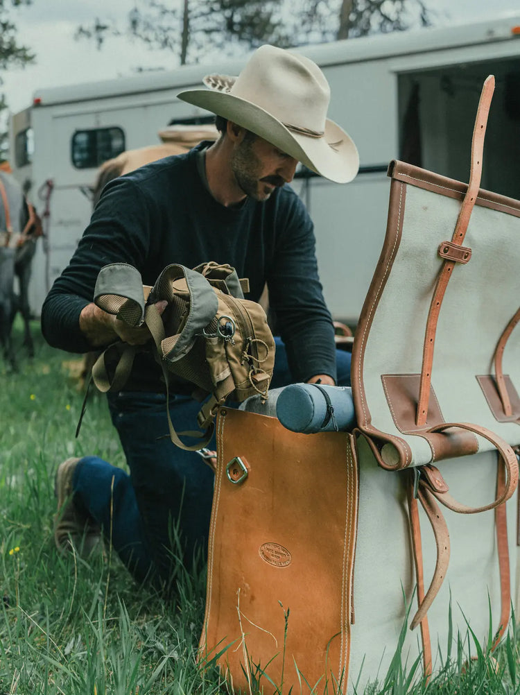Man kneeling down, placing items inside a pack saddle pannier that is lying in the grass, with a horse trailer in the background