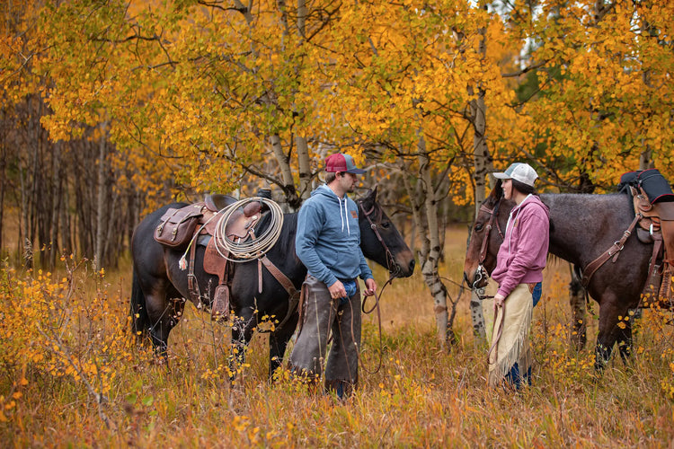 A man and a woman wearing Outfitters Supply Logo hoodies and ball caps, standing in a fall setting with their horses
