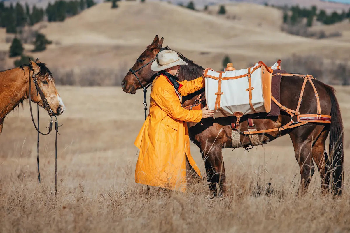 man wearing a rain slicker, adjusting canvas and leather pannier on pack saddle on a horse