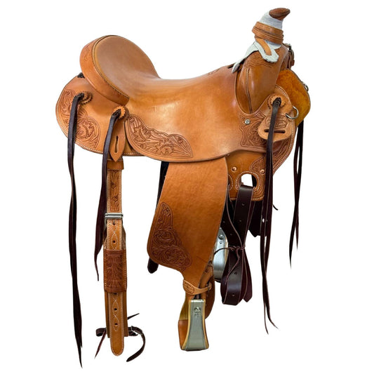 A classic brown leather western saddle with intricate hand-tooled floral patterns, black leather straps, and silver metal hardware, isolated on a white background.