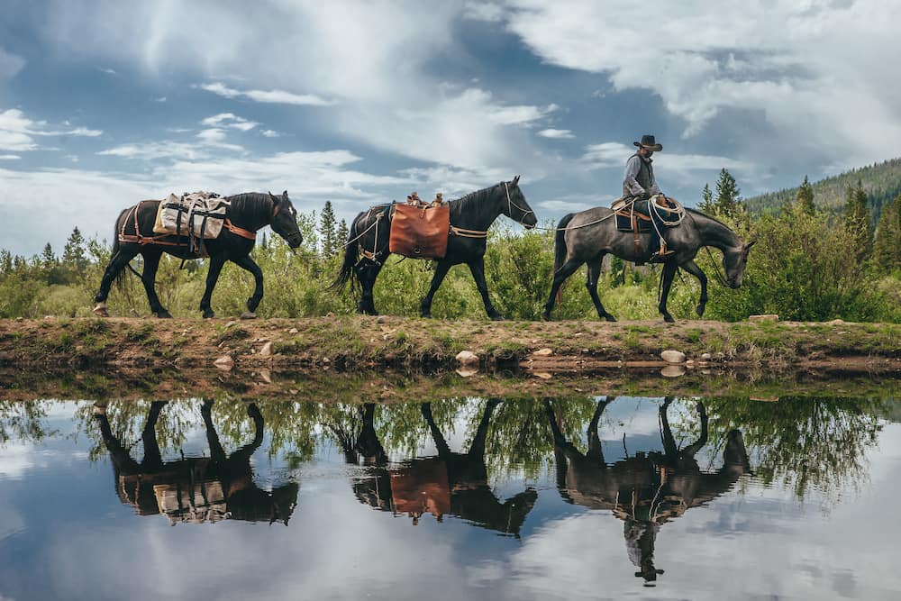 Three horses with riders walking along a path by a reflective water body with a scenic background.