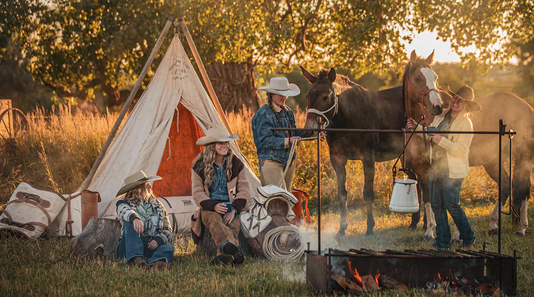 Group of people with horses around a campfire and tent in a natural setting
